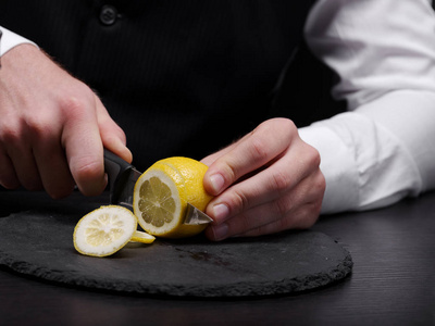 s hands cutting a ripe, raw, fresh yellow lemon on a black cutti