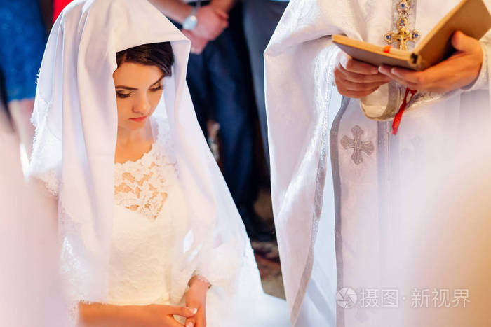 美丽的黑发新娘穿着白色婚纱在披肩下祈祷牧师在基督教教堂举行仪式