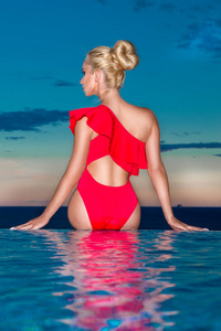 美丽的金发女人模型与令人惊叹的身体站在池的优雅的红色泳衣和背景是惊人视图的海和天空