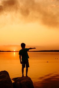 小男孩发射纸船只从河边。在远方的浮船。美丽的夕阳。折纸。河里。湖