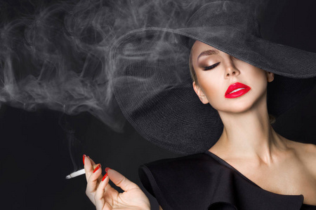 优雅的女人,黑党徽与香烟在手拿着帽子.在黑色背景上照片