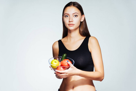 年轻的女运动手表她的身影，在她手里捧着一盘有用的蔬菜和水果