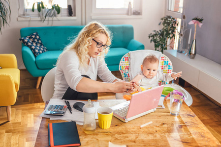 妈妈带着眼镜和宝宝坐在办公桌前，在家里的办公室里用智能手机