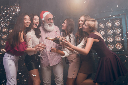 一个大胡子的圣诞老人的帽子到女孩的眼镜，笑容倒香槟。现代圣诞老人在圣诞帽子有女孩玩的俱乐部和纸屑。2018 年新的一年快乐