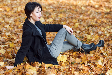女孩穿件黑色的大衣，坐在黄色的叶子，金秋时节，城市公园