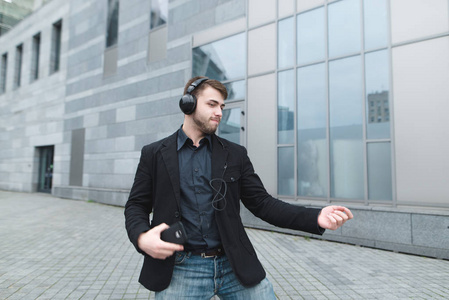 一个积极的人穿着夹克在城市景观的背景下, 用耳机跳舞。快乐的积极的商人是高兴
