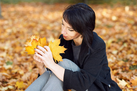 女孩穿件黑色的大衣，坐在黄色叶子在秋天的城市公园，秋天的季节