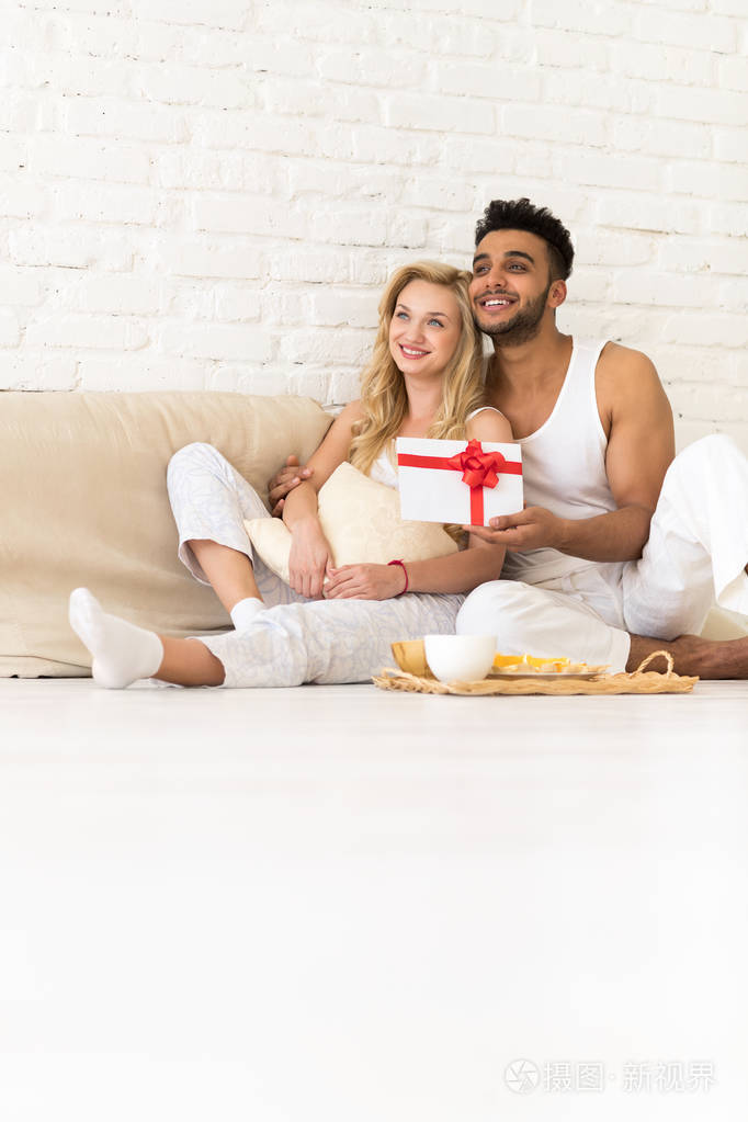 年轻夫妇坐在枕头地板上, 快乐的微笑拉美裔男子妇女举行礼物信封与丝带恋人在卧室