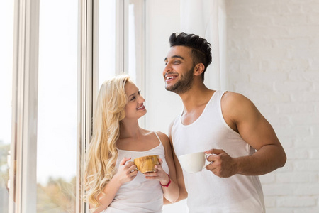年轻漂亮的夫妇站在大窗口附近, 喝早茶咖啡杯, 快乐的微笑拉美裔男人女人
