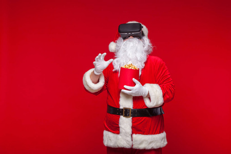 圣诞老人穿着虚拟现实的护目镜和一个红色的水桶与爆米花, 在红色的背景。圣诞节