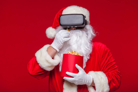 圣诞老人穿着虚拟现实的护目镜和一个红色的水桶与爆米花, 在红色的背景。圣诞节