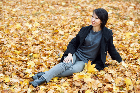 女孩穿件黑色的大衣，坐在黄色叶子在秋天的城市公园，秋天的季节