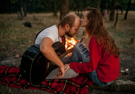 迷恋夫妇坐在野餐的森林与吉他的篝火火焰背景