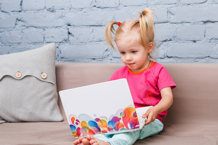 儿童女孩三岁的使用培训膝上型电脑坐在沙发上的蓝色砖墙室内。一个蓝色的眼睛和她的头上有滑稽的尾巴的金发碧眼的婴儿穿着明亮的 jer