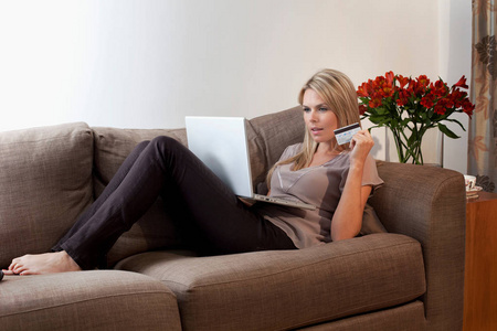 有魅力的女人躺在沙发上，拿着笔记本电脑和信用卡。网上购物概念
