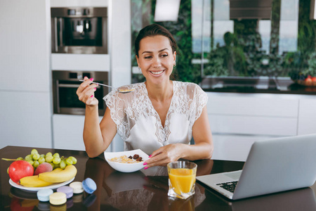 美丽的幸福的女人的肖像工作与笔记本电脑, 而早餐与谷物和牛奶。在家吃饭