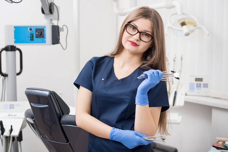 在现代牙科诊所里手持牙科器械的年轻友好牙医的肖像。医生戴着眼镜, 蓝色制服, 蓝色手套。牙科