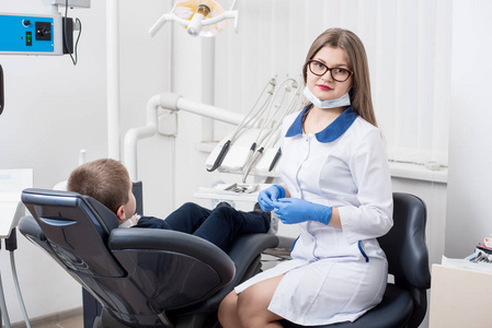 女牙医检查和工作的男孩病人。在牙医的椅子上的孩子男孩。医生看着相机。牙医办公室