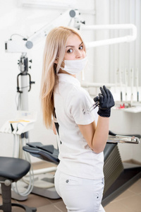 在现代牙科诊所里手持牙科设备的年轻女牙医的肖像。医生戴口罩, 黑色手套, 并期待在相机。牙科