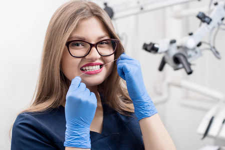 近距离的肖像美丽的女牙医与牙线在牙科办公室。医生身穿蓝色制服和手套, 戴眼镜。牙科.口腔设备的背景