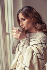 年轻漂亮的女人站在窗户旁边, 裹着毯子, 喝着热咖啡。舒适.秋天.温馨温馨的夜晚