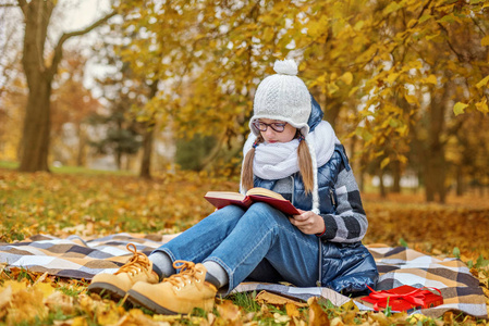 戴眼镜的少年学生女孩在公园里学习教科书坐在一个舒适的白色围巾和帽子的格子