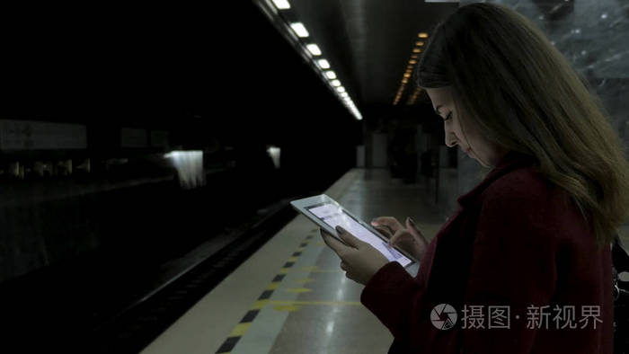 穿红大衣的女孩在地铁站使用智能手机或平板电脑, 等待火车。妇女使用手机和站在城市地铁史丹顿