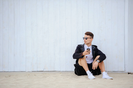 左侧脸时髦的男人在白色的墙上坐在沙滩上, 看着左边, 穿着衬衫和短裤