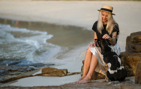 漂亮的金发女郎, 戴着草帽, 坐在岩石上, 抚摸着好狗狗。相同的颜色。泰国海滩。晨走
