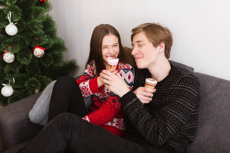 年轻快乐的情侣坐在沙发上的肖像在家里, 愉快地花时间一起吃冰淇淋与圣诞树附近