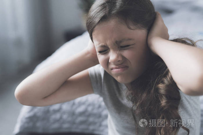 黑头发的女孩坐在床旁边皱着眉头, 用她的双手捂住耳朵
