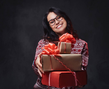 一位微笑的黑发女性，穿着红色套头衫和眼镜，在灰色背景下持有圣诞礼物。