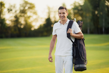 一个穿着白色西装的男人带着一个高尔夫球棍袋走在高尔夫球场周围, 微笑着