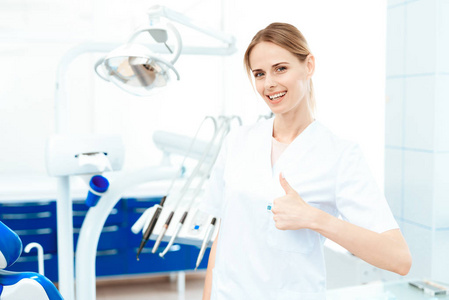 女牙医在牙科诊所的牙科设备背景下摆姿势。她竖起大拇指