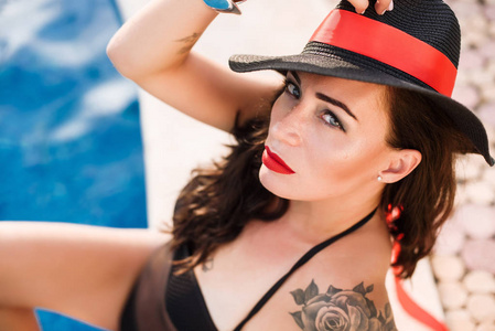 年轻的西班牙时尚女性，戴着黑色帽子和泳衣，红唇在游泳池摆姿势。 珠宝纹身明亮的化妆。 时尚风格。 夏天和假期的概念。