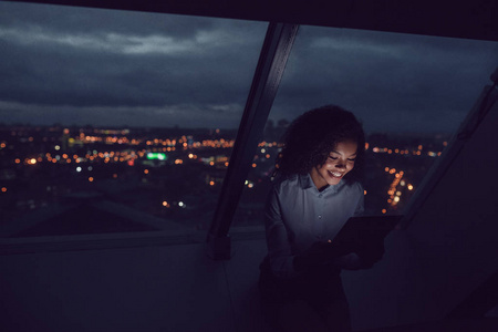 商业妇女使用一个平板电脑坐在办公室的窗口在一个晚上城市的背景