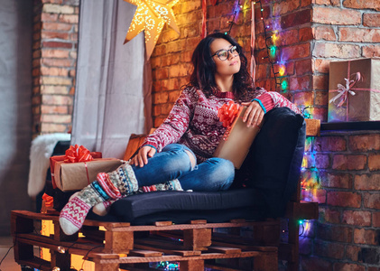 全身形象的深色女性眼镜，穿着牛仔裤和红色毛衣，摆在一个木制沙发上的房间与圣诞节装饰。
