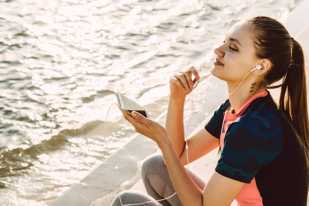 穿着运动服的女孩坐在河岸上看着水, 听着耳机里的音乐