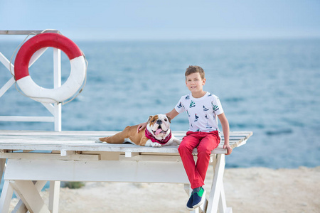 英俊的男孩青少年 happyly 花时间与他的朋友斗牛犬在海边的孩子狗控股玩两个海星接近生活浮标穿红色长裤拖鞋和 tshirt