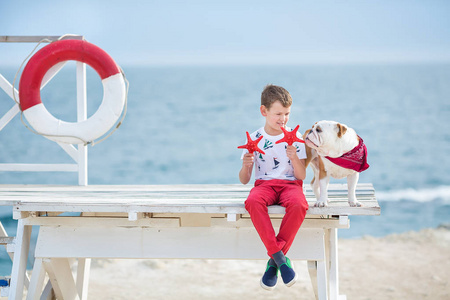 英俊的男孩青少年 happyly 花时间与他的朋友斗牛犬在海边的孩子狗控股玩两个海星接近生活浮标穿红色长裤拖鞋和 tshirt