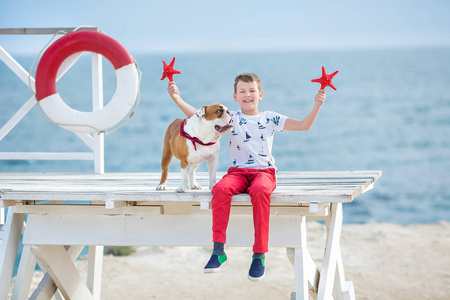 英俊的男孩青少年 happyly 花时间与他的朋友斗牛犬在海边的孩子狗控股玩两个海星接近生活浮标穿红色长裤拖鞋和 tshirt