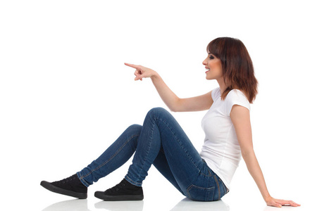 微笑的年轻女子坐在地板上, 指着看着远处.侧面视图照片