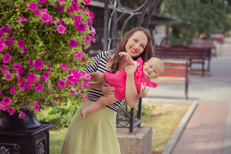 图片的年轻女子黑发母亲在柔软的斑马衬衫和象牙色的裙子快乐地玩与女婴在城市街道公园接近粉红色玫瑰花夏季时间孩子