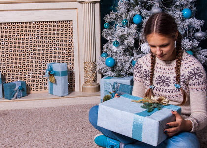 愉快的女孩举行圣诞节礼物箱子在美丽的房间内部的假日早晨。手持圣诞礼物的女孩子在圣诞树和壁炉的装饰附近。寒假概念