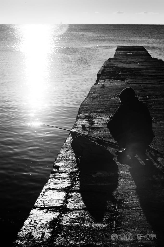 渔夫在海墩上黑白捕鱼黎明时的秋天海景背光中有钓鱼竿的人的轮廓