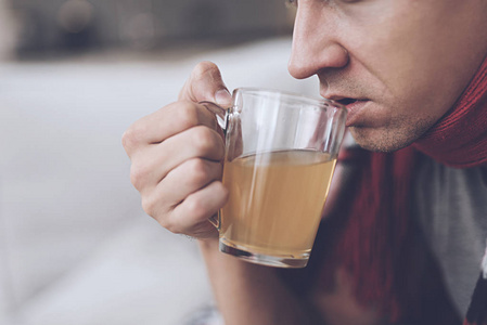 一个感冒的人坐在沙发上, 躲在红地毯后面。他喝药用茶