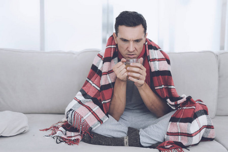 一个感冒的人坐在沙发上, 躲在红地毯后面。他喝药用茶