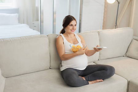 一个孕妇坐在家里的轻便沙发上。她拿着一瓶薯片和一台遥控器