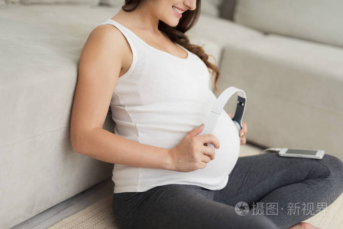 一个孕妇坐在家里的浅色地板上她把耳机放在肚子上