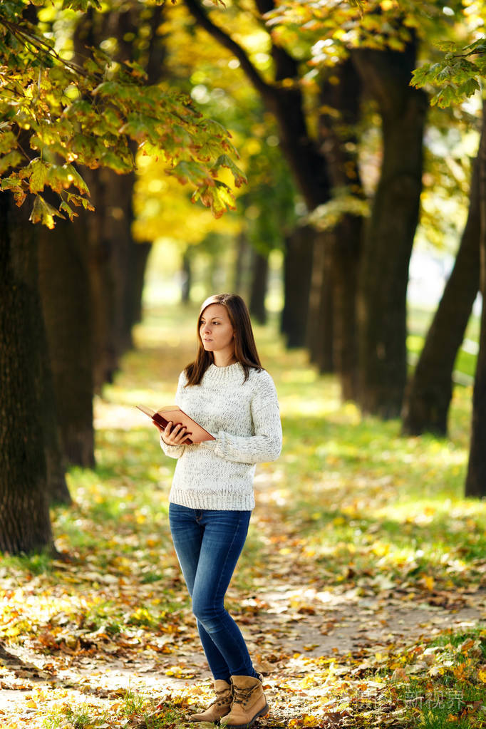 在一个温暖的日子里, 一个穿着白色毛衣的漂亮笑脸棕色头发的女人站在秋天城市公园里的一本红色的书。秋天金黄叶子。阅读理念